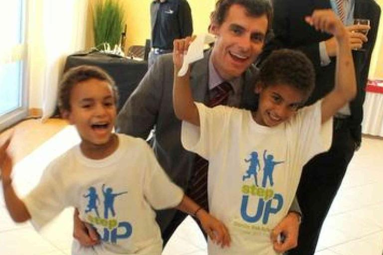 A man in a suit poses with two smiling children wearing matching "Step Up" t-shirts, all three appearing happy at an indoor event.