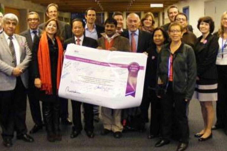 A group of people pose indoors, holding a large signed poster with text and graphics, while others stand around them, facing the camera.