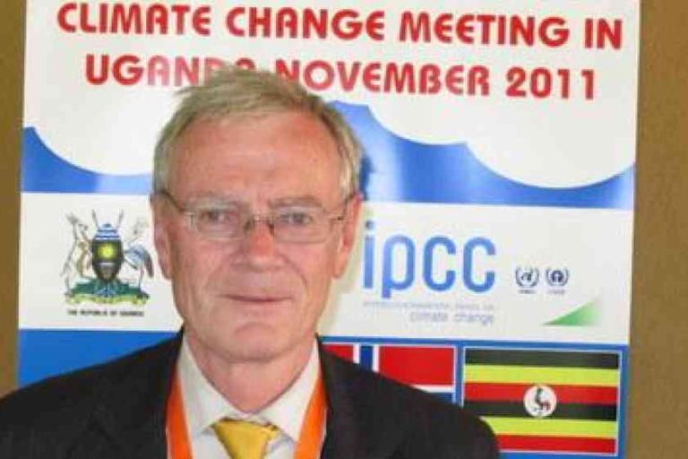 A man stands in front of a poster for a climate change meeting in Uganda, dated November 2011, with the IPCC logo and various flags displayed.