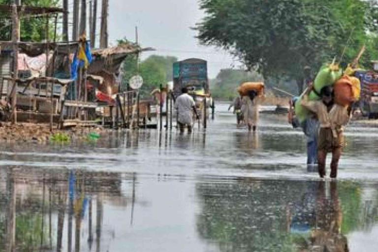 People walk through a flooded street lined with makeshift structures and trees, carrying belongings on their heads and shoulders.
