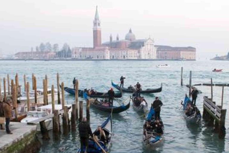 Gondolas with passengers navigate a canal in Venice, Italy, with the historic San Giorgio Maggiore island and church visible in the background.