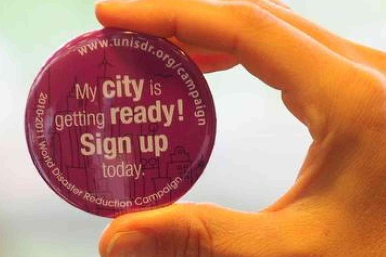 Hand holding a round purple button that reads, "My city is getting ready! Sign up today." and includes a website URL and campaign details.