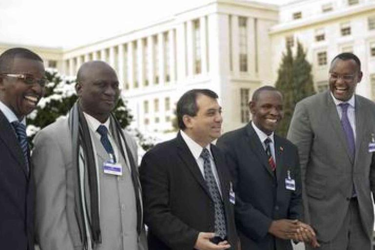 Five men in suits and conference badges stand outdoors in front of a large building, smiling and conversing on a winter day.