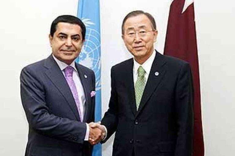 Two men in business attire shake hands in front of a United Nations flag and another flag in an official setting.