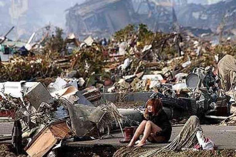 A person sits amid extensive debris and destruction, surrounded by rubble and damaged structures after a natural disaster.