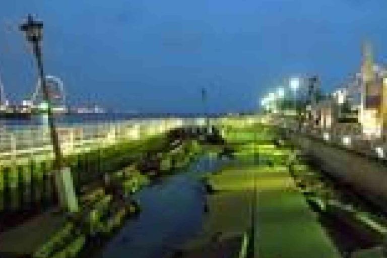 A seaside promenade at dusk with green-lit walkway, streetlights, buildings on the right, and a body of water on the left.