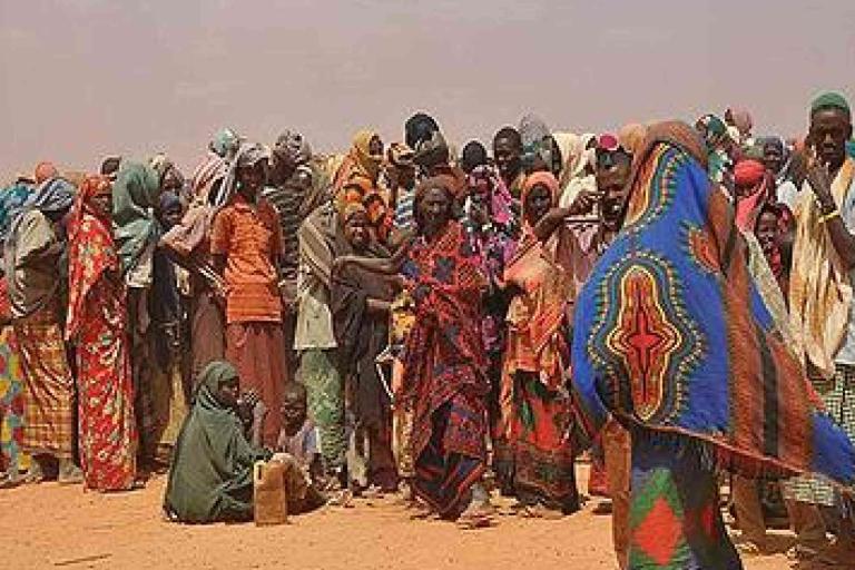 A large group of people, including adults and children, stand and sit closely together outdoors in a dry, dusty environment. Many wear colorful patterned clothing and head coverings.