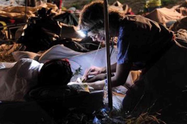A person wearing a headlamp provides medical care to someone lying on a cot outdoors at night, surrounded by other patients and emergency supplies.