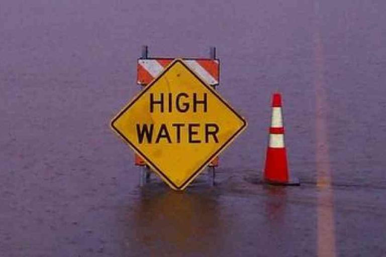 Yellow "HIGH WATER" warning sign and orange traffic cone partially submerged in floodwater.
