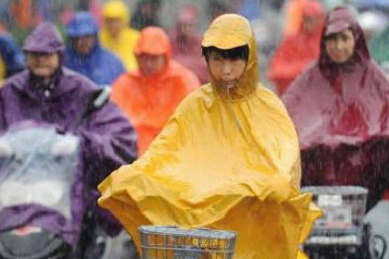 People wearing colorful rain ponchos ride bicycles and scooters in the rain, with water droplets visible in the air.