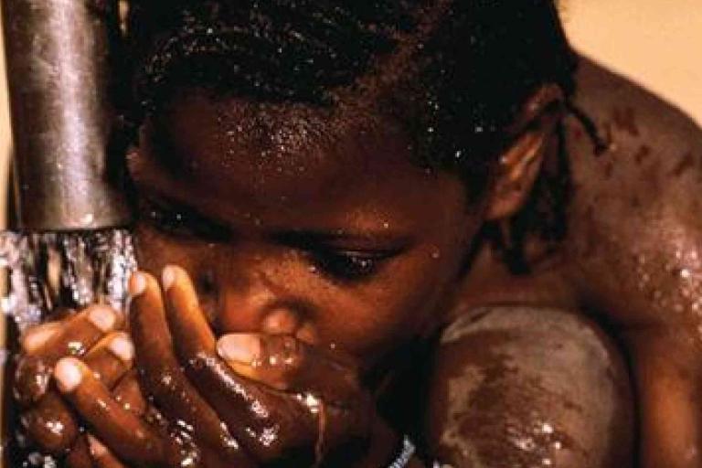 A child kneels and drinks water with cupped hands from a pipe, with droplets visible on their face and arms.