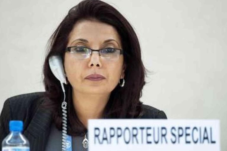 A woman wearing glasses and a headset sits at a desk with a sign reading "Rapporteur Special.