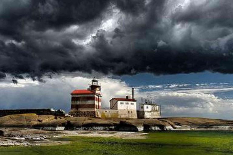 A lighthouse and adjacent buildings stand on a rocky shore under a dramatic, dark, and cloudy sky, with green water in the foreground.