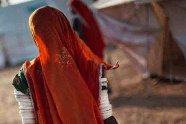 Person wearing an orange headscarf and patterned clothing stands outdoors, facing away, with tents and another person visible in the background.