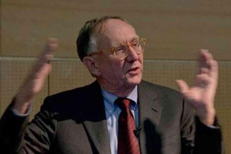 An older man in a suit and glasses gestures with his hands while speaking in a lecture or presentation setting.