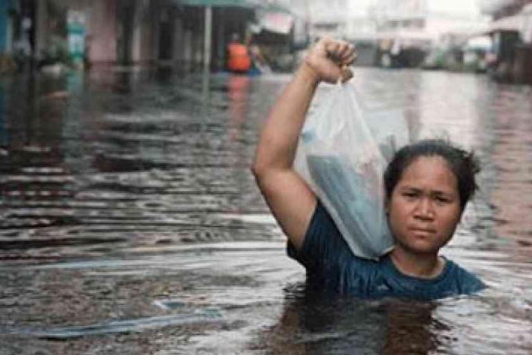 A woman wades through chest-deep floodwater on a city street, holding a plastic bag above her head to keep it dry.