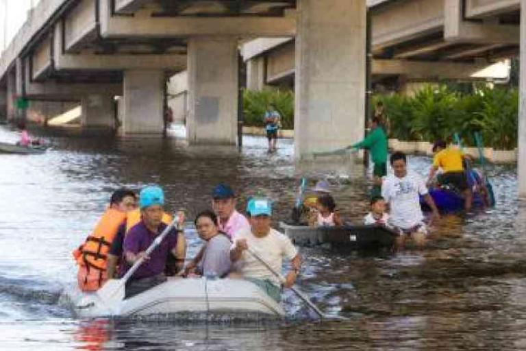 People ride boats through a flooded street under a bridge, navigating high water with oars. Others are seen wading or floating in the background.