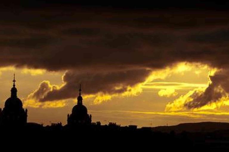 Dark clouds partially obscure a golden sunset sky, with the silhouette of two domed towers and city rooftops visible in the foreground.