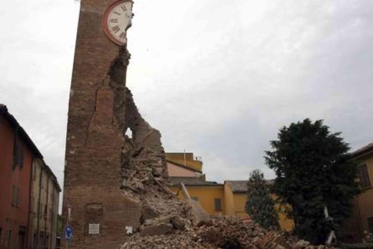 A partially collapsed brick clock tower stands amid rubble between buildings, showing significant earthquake damage under an overcast sky.