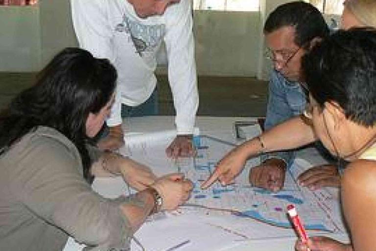Five people discuss and point at a large map or blueprint spread out on a table during a meeting.