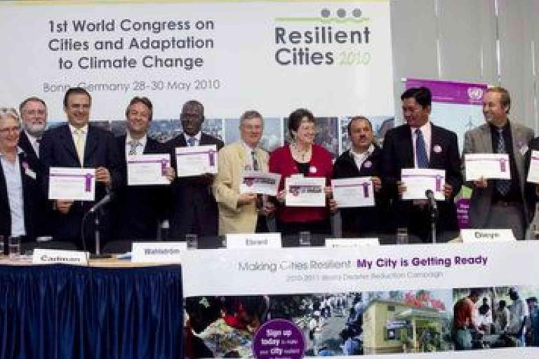 A group of people stand and sit at a table holding certificates at the 2010 Resilient Cities Congress in Bonn, Germany, with event banners displayed behind them.