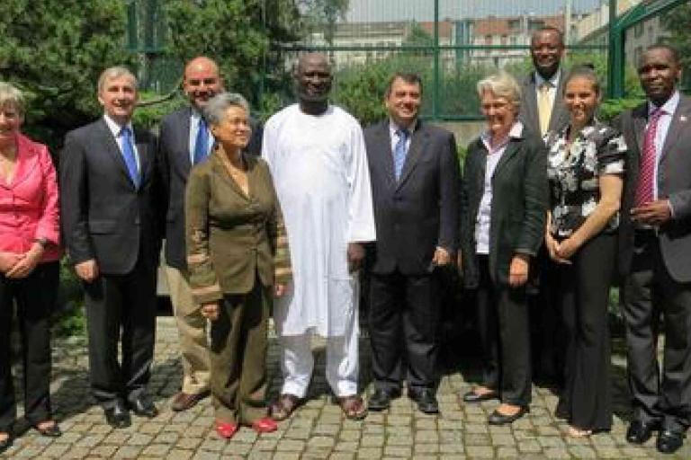 Ten people, dressed in business or formal attire, stand in a line outdoors on a stone path, posing for a group photo with trees and a building in the background.