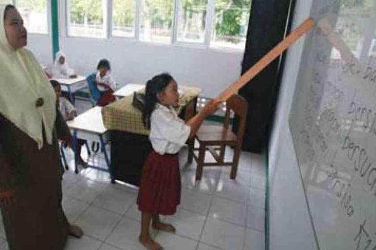 A girl in a school uniform points to words on a whiteboard with a stick while a teacher observes; other students are seated at desks in the classroom.