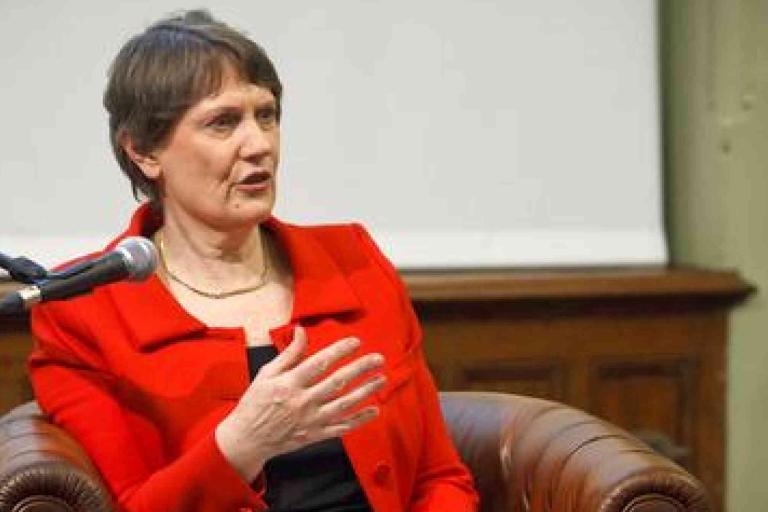 A person in a red jacket sits in a brown leather chair, speaking into a microphone with one hand raised, against a blurred indoor background.