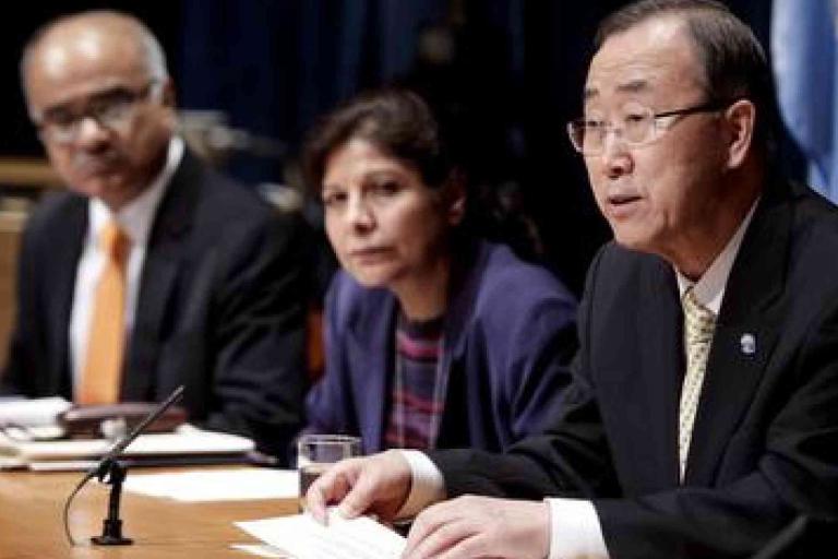 Three people sit at a table during a formal meeting; one man speaks into a microphone while the others listen attentively.