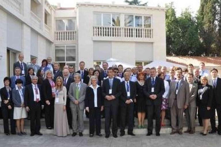 A large group of people in formal attire pose for a photo outside a building with a balcony and trees in the background.