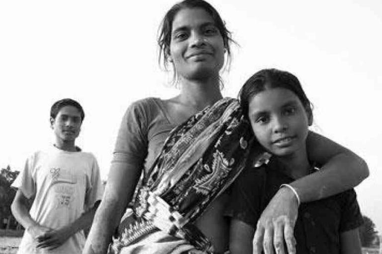 A woman stands with her arm around a young girl, both looking at the camera, while a man stands in the background outdoors in a black-and-white photo.