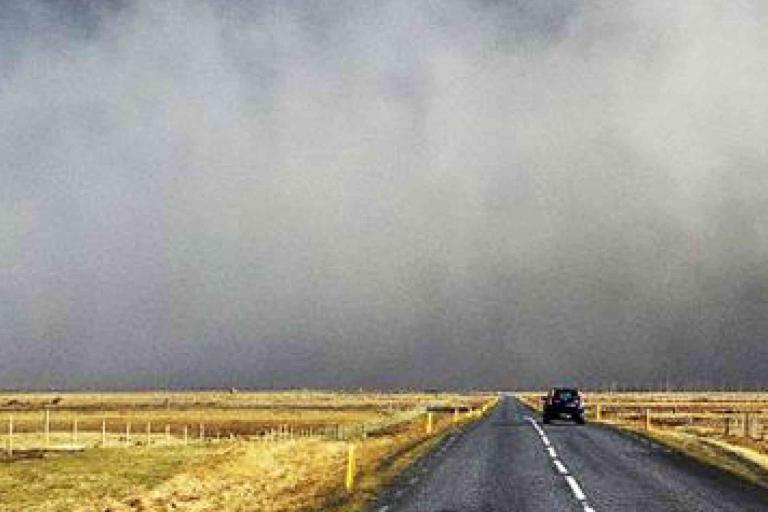 A car drives down a rural two-lane road with fields on either side under a sky filled with thick, gray fog or dust.