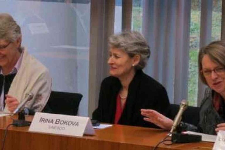 Three women sit at a conference table with microphones and nameplates, including one labeled "Irina Bokova UNESCO," participating in a formal discussion.