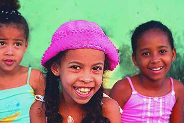 Three young girls sit together in front of a green wall; the girl in the center wears a pink hat and smiles widely.