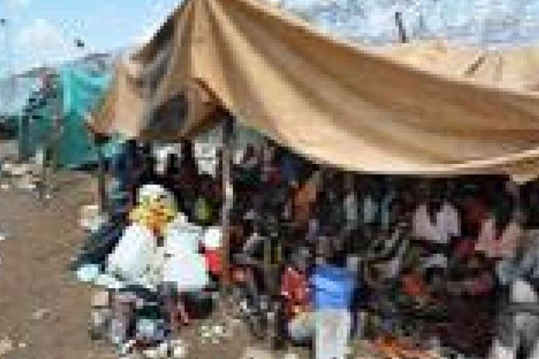A group of people sit and stand under a large makeshift tent in a crowded outdoor area with scattered debris.