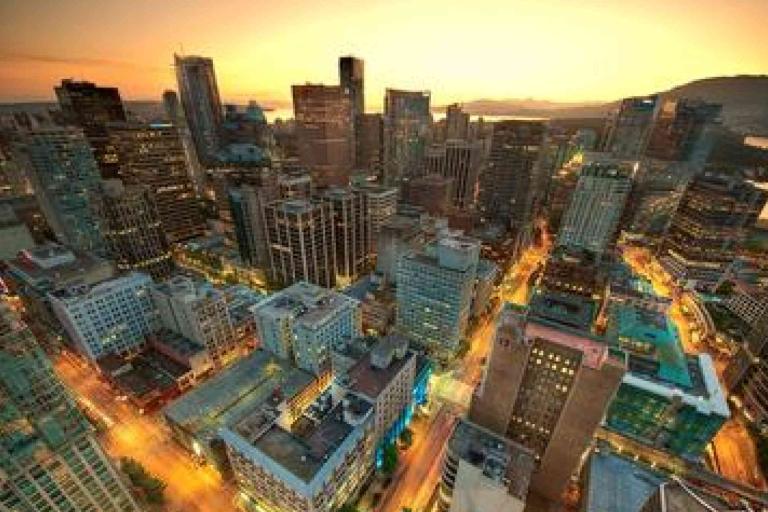 Aerial view of a city skyline at sunset, showing tall buildings, illuminated streets, and a body of water in the background.