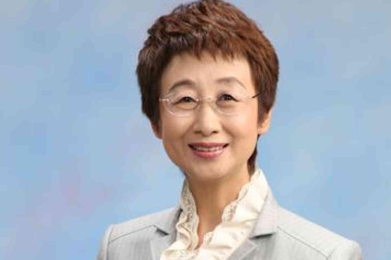 A woman with short brown hair, glasses, and a light gray blazer over a ruffled white blouse, smiling in front of a plain blue background.