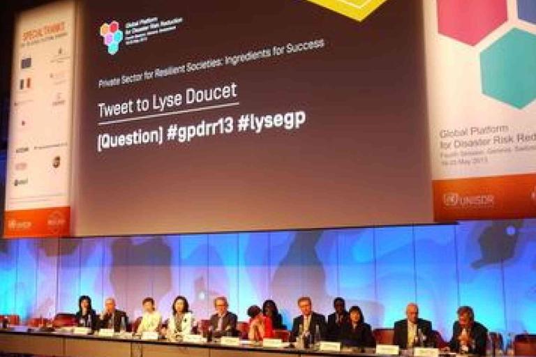 A panel of speakers sits at a long table on stage at the Global Platform for Disaster Risk Reduction, with a large screen displaying event information behind them.