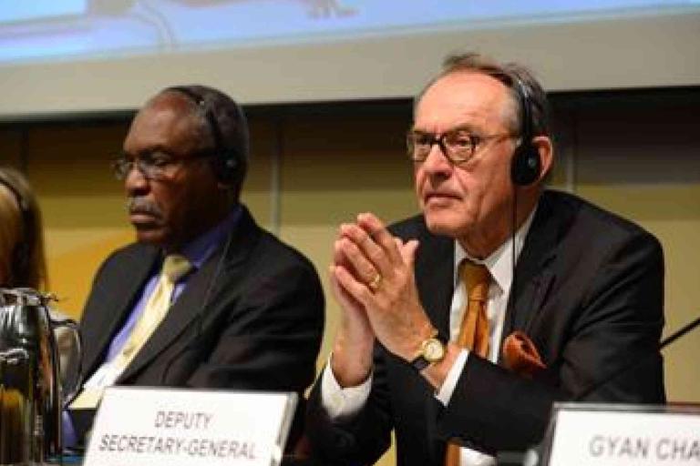 Three people sit at a conference table wearing headsets; the man in front is labeled as the Deputy Secretary-General.