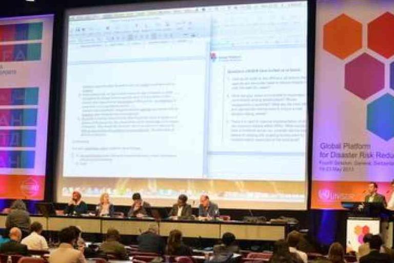 A conference panel sits at a long table facing an audience, with large presentation slides projected on screens behind them.