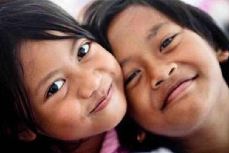 Two children smiling closely together, with their heads touching and looking directly at the camera.