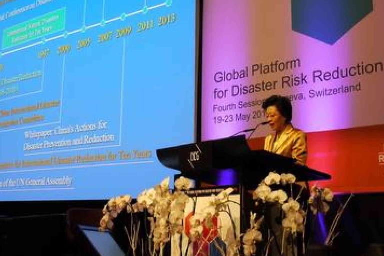 A woman speaks at a podium during the Global Platform for Disaster Risk Reduction conference in Geneva, with a presentation slide projected behind her.