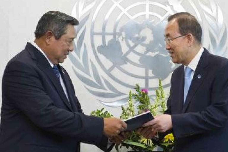 Two men in suits exchange a document in front of a United Nations emblem, with flowers and a flag in the background.