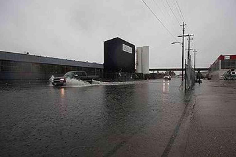 Vehicles drive through a flooded industrial street on a rainy day, with water covering most of the road and overcast skies overhead.