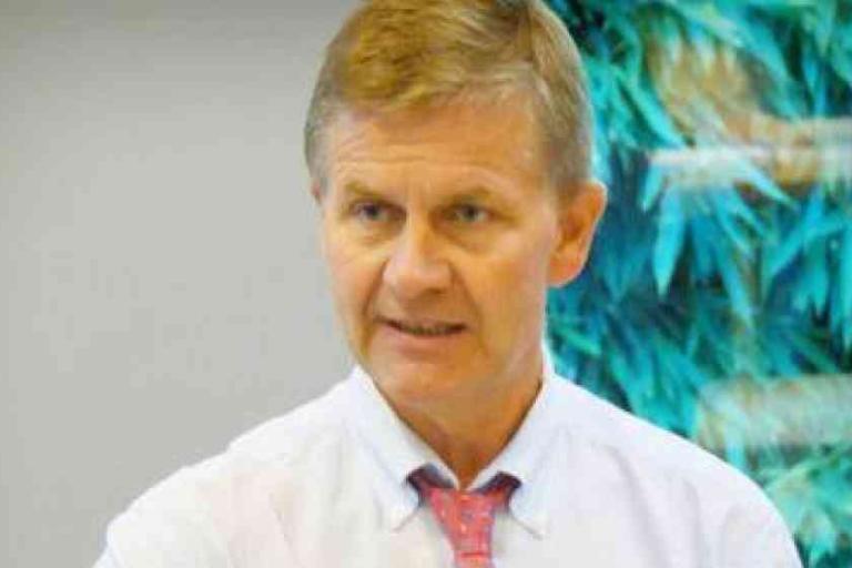 A man in a white shirt and red tie is speaking indoors, with a blurred, leafy background behind him.