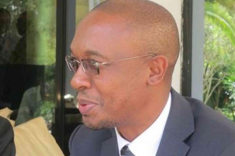 A man in a suit and glasses sits indoors, facing left, with greenery visible outside through a window behind him.