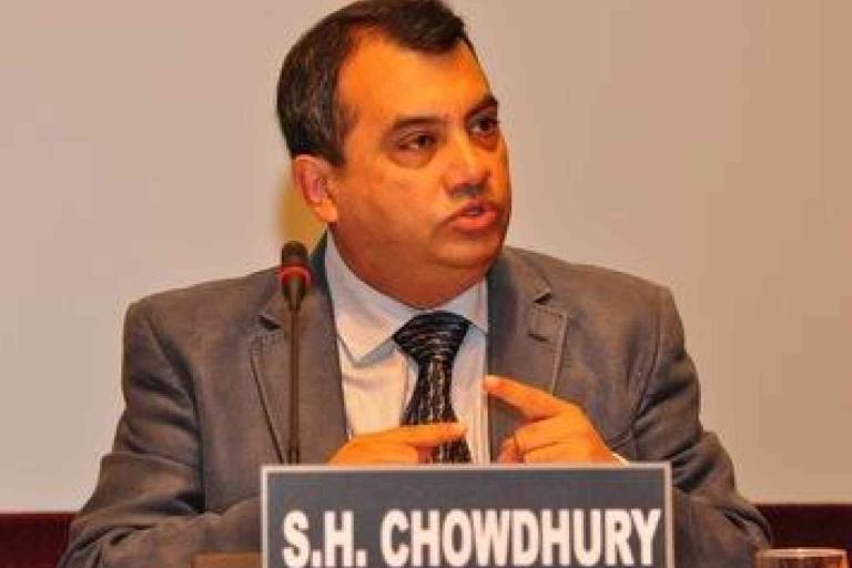 A man in a suit speaking at a microphone, seated behind a nameplate labeled "S.H. CHOWDHURY.
