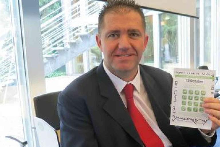 A man in a suit and red tie sits indoors, holding up a calendar or planner open to the month of October.