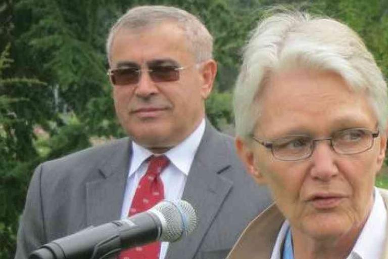 Two older adults, a man in a suit and a woman with glasses, stand outdoors near a microphone with trees in the background.