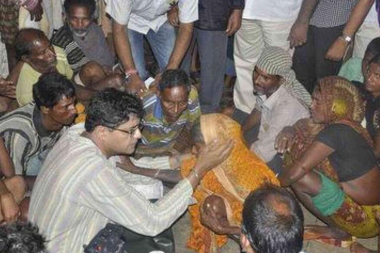 A man in glasses talks to and comforts a woman in a yellow sari, surrounded by a group of seated and standing people in an indoor setting.